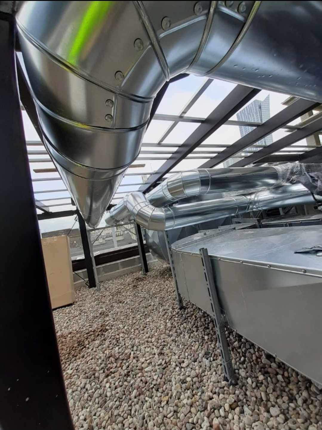 Srebrne, metalowe rury wentylacyjne na dachu budynku, widoczne z dołu, z elementami konstrukcyjnymi i kamienistym podłożem.