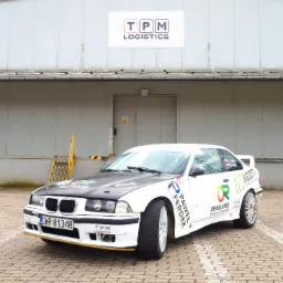 Biały samochód sportowy z naklejkami sponsorskimi na tle magazynu z banerem TPM Logistics