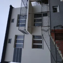 Dwupiętrowy dom z białą elewacją, balkonami z ozdobnymi metalowymi balustradami i zewnętrznymi schodami z ceglanym obramowaniem.