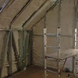 Poddasze z metalową konstrukcją i izolacją natryskową z pianki, widoczny fragment podłogi i drabina z platformą roboczą.