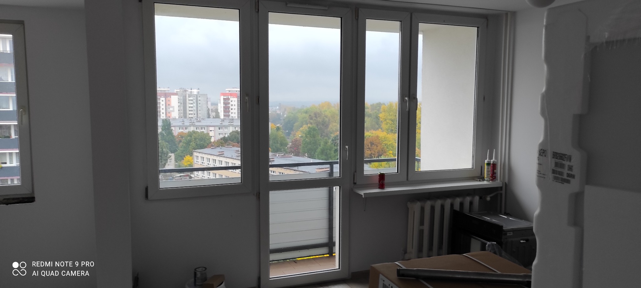 Wnętrze mieszkania w trakcie remontu: widok z okna na miasto, częściowo pomalowane ściany, materiały budowlane i narzędzia na podłodze, grzejnik pod parapetem.