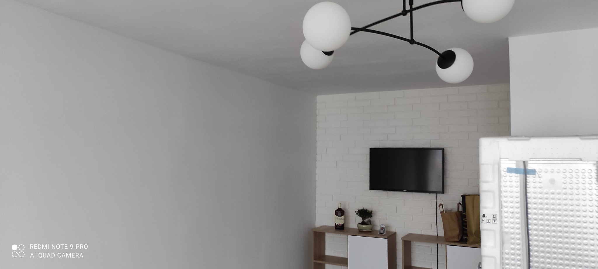 Wnętrze pokoju z białą ceglaną ścianą, nowoczesną lampą sufitową z kulistymi kloszami, telewizorem i szafką RTV. Na pierwszym planie fragment opakowania z widoczną perforacją.