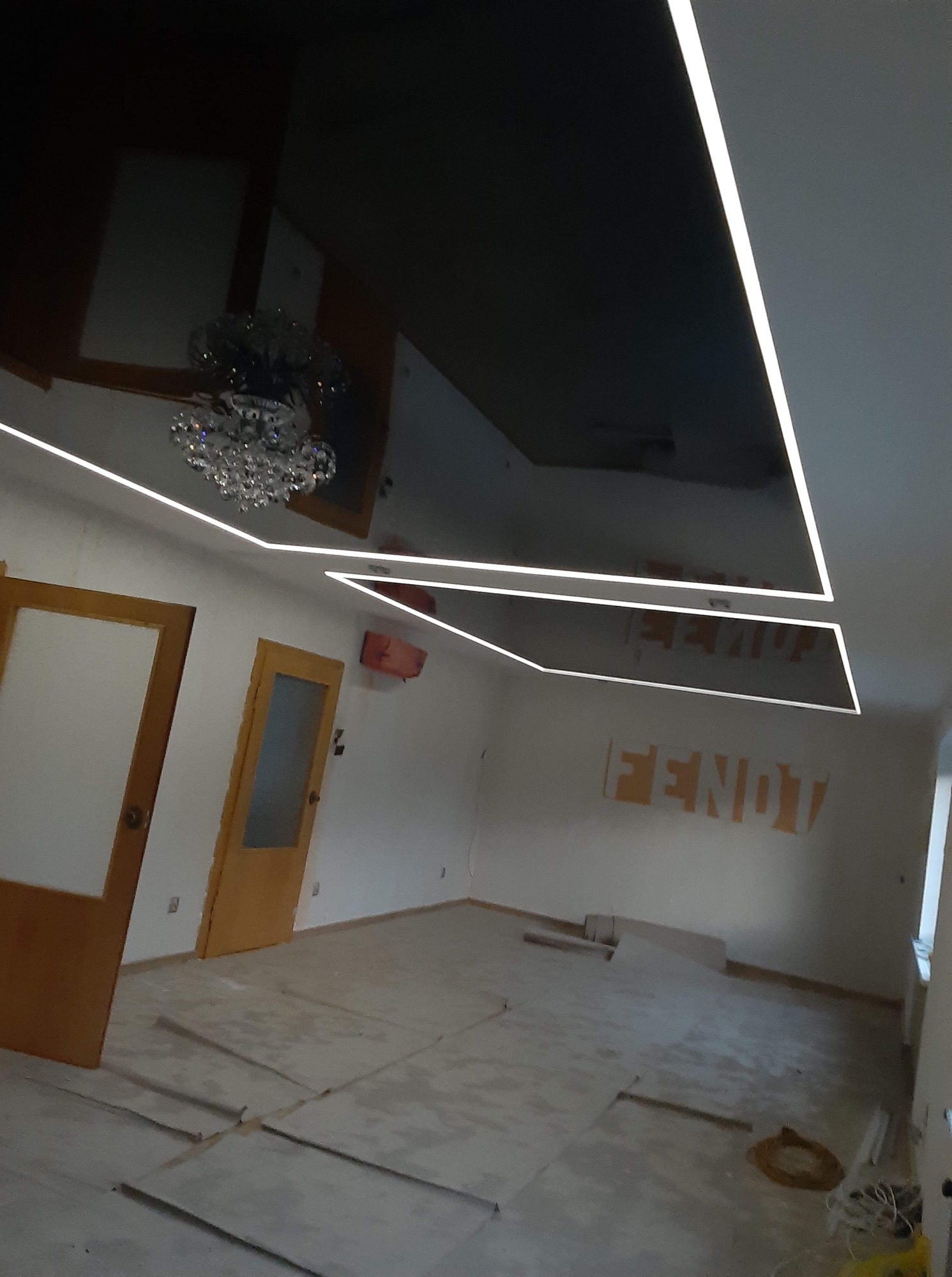 Czarne lustrzane wykończenie sufitu z oświetleniem LED w kształcie geometrycznym, odbijające kryształowy żyrandol w remonowanym pomieszczeniu z odsłoniętymi ścianami.