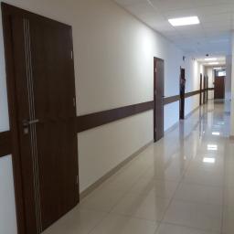 Okna Ola-sprzedaż, montaż drzwi, okien i bram garażowych - Długi korytarz biurowy z rzędem ciemnobrązowych drzwi z metalowymi klamkami i trzema pionowymi, srebrnymi paskami, zabezpieczony brązową listwą na ścianie, wyłożony błyszczącymi, beżowymi płytkami.