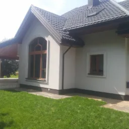 Fragment elewacji domu z oknem łukowym i mniejszym oknem prostokątnym, ciemna dachówka, świeżo skoszony trawnik.