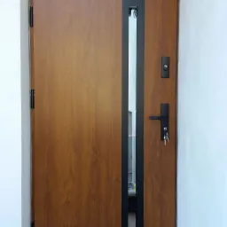 Brązowe drzwi wejściowe z czarnym pionowym panelem i szybą, nadświetle okienne, widoczne klucze w zamku, białe ściany.