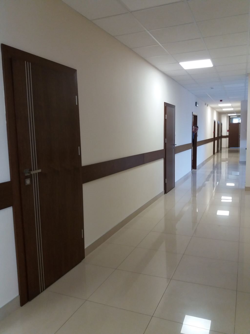 Długi korytarz biurowy z rzędem ciemnobrązowych drzwi z metalowymi klamkami i trzema pionowymi, srebrnymi paskami, zabezpieczony brązową listwą na ścianie, wyłożony błyszczącymi, beżowymi płytkami.