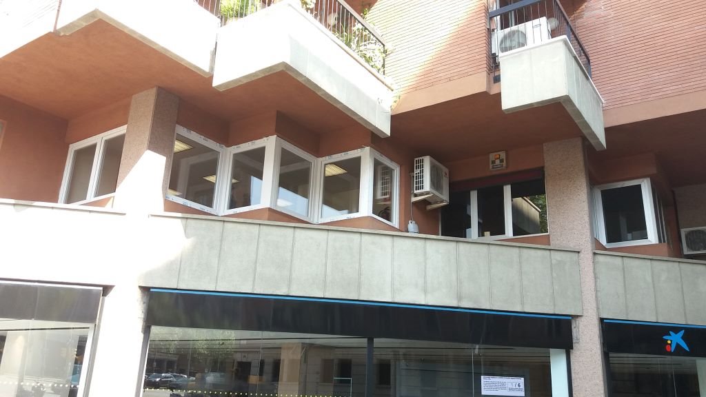 Elewacja budynku z zamontowanymi białymi oknami plastikowymi, widoczne balkony i klimatyzatory, dolna część budynku to witryna sklepowa z logo banku.