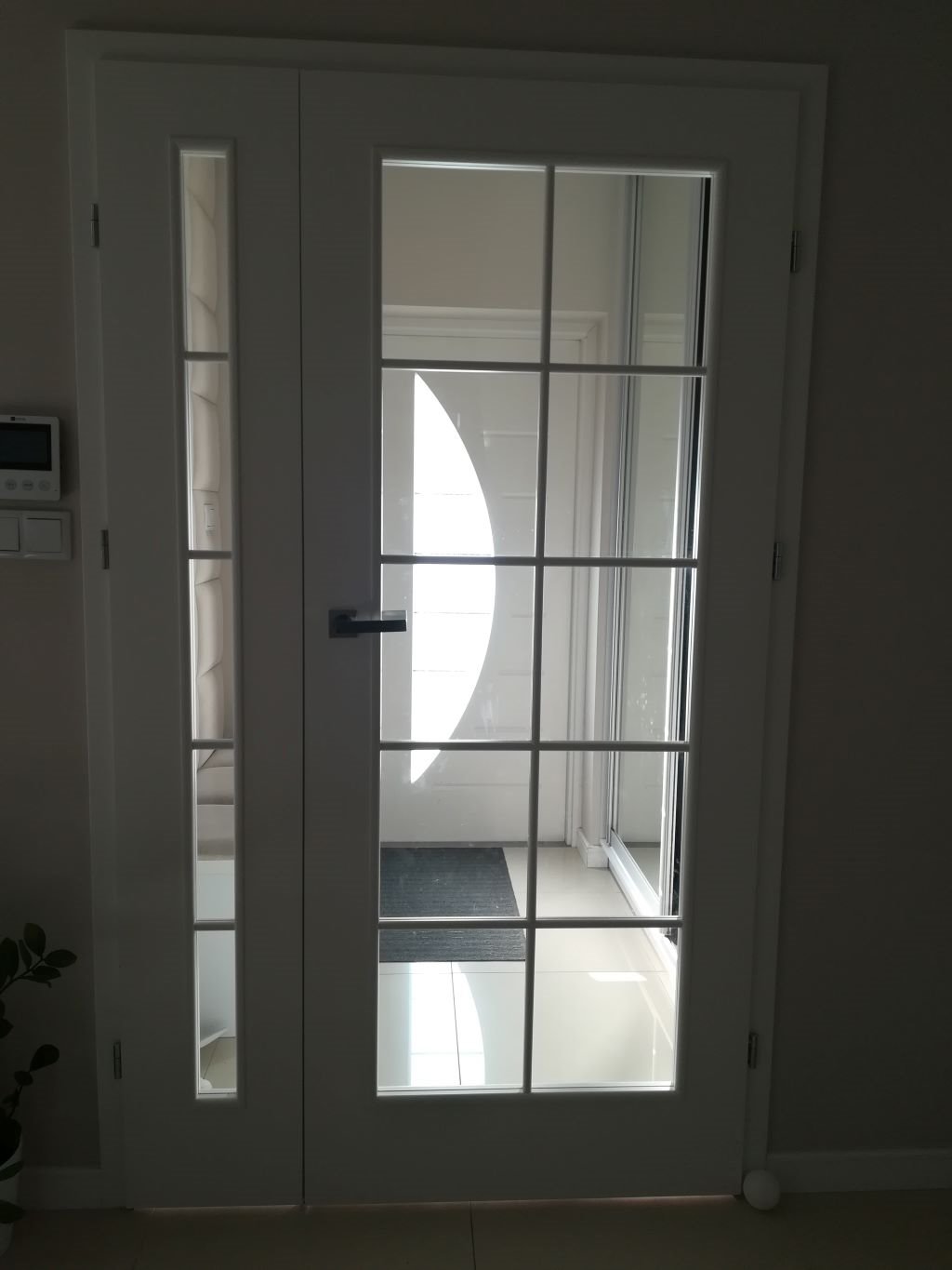 Białe, przeszklone drzwi wewnętrzne z czarną klamką, widok na jasne pomieszczenie z jasną podłogą i czarną wycieraczką, z boku widoczny fragment ściany z włącznikiem.