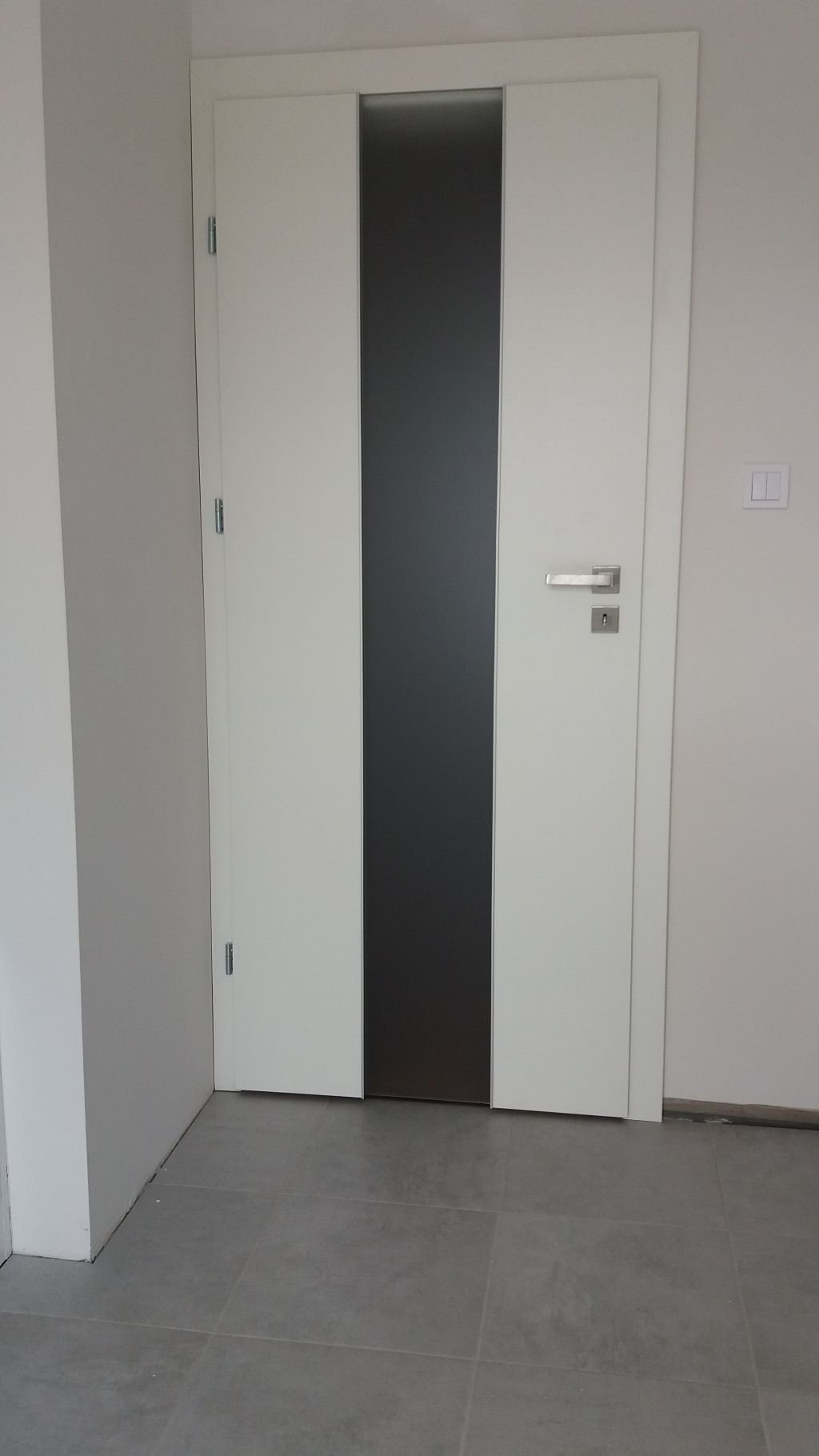 Nowoczesne białe drzwi wewnętrzne z pionowym, szarym panelem szklanym i srebrną klamką, widoczne na tle szarej podłogi z płytek.