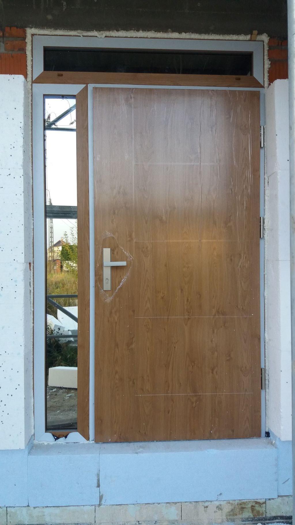 Brązowe drzwi wejściowe z klamką, częściowo otwarte, w surowym otworze murarskim z widoczną izolacją i folią ochronną na powierzchni.