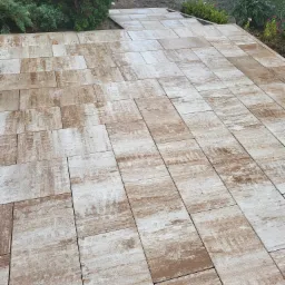 Ułożona ścieżka z prostokątnych kostek brukowych w odcieniach beżu i brązu, prowadząca przez ogród z elementami zieleni i placem zabaw w tle.