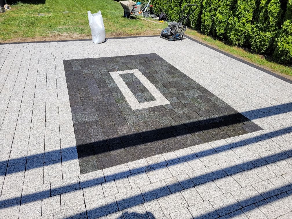 Ułożona szara kostka brukowa z dekoracyjnym wzorem w odcieniach szarości i białym prostokątem pośrodku, widoczna zagęszczarka w tle.