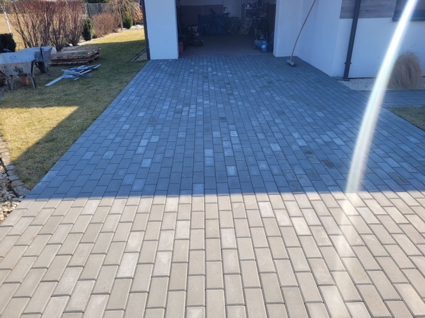 Ułożona szara kostka brukowa na podjeździe do garażu, widoczny fragment trawnika z taczką i narzędziami.