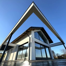 B. M. GROUP Marek Bala - Nowoczesny dom z białą elewacją i dużymi oknami, widziany z dołu pod kątem, z elementami konstrukcyjnymi w kształcie litery V na tle błękitnego nieba.