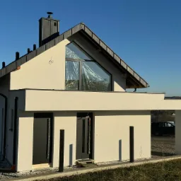 Nowoczesny dom jednorodzinny z jasną elewacją i antracytową stolarką okienną, częściowo osłoniętą folią ochronną podczas prac wykończeniowych, na tle błękitnego nieba.