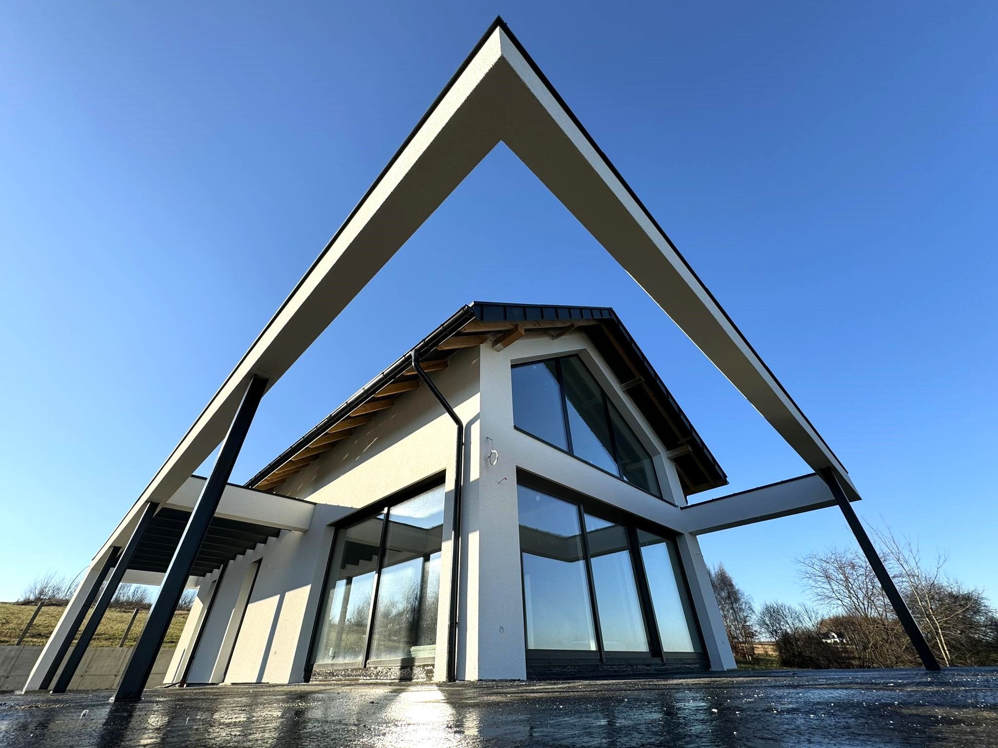 Nowoczesny dom z białą elewacją i dużymi oknami, widziany z dołu pod kątem, z elementami konstrukcyjnymi w kształcie litery V na tle błękitnego nieba.