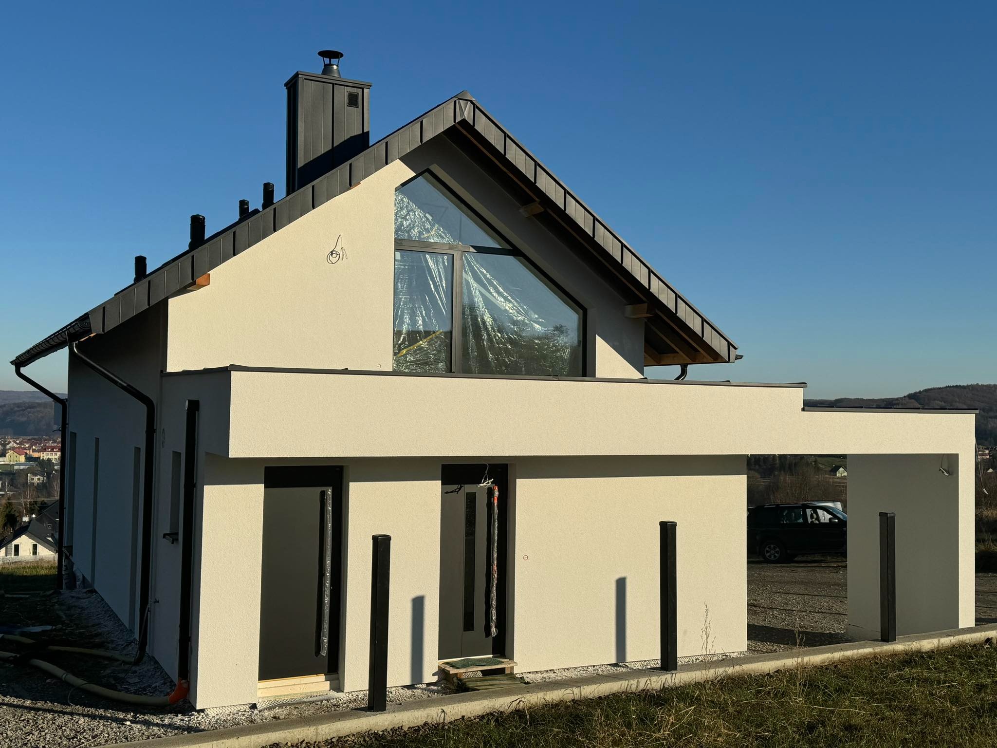 Nowoczesny dom jednorodzinny z jasną elewacją i antracytową stolarką okienną, częściowo osłoniętą folią ochronną podczas prac wykończeniowych, na tle błękitnego nieba.