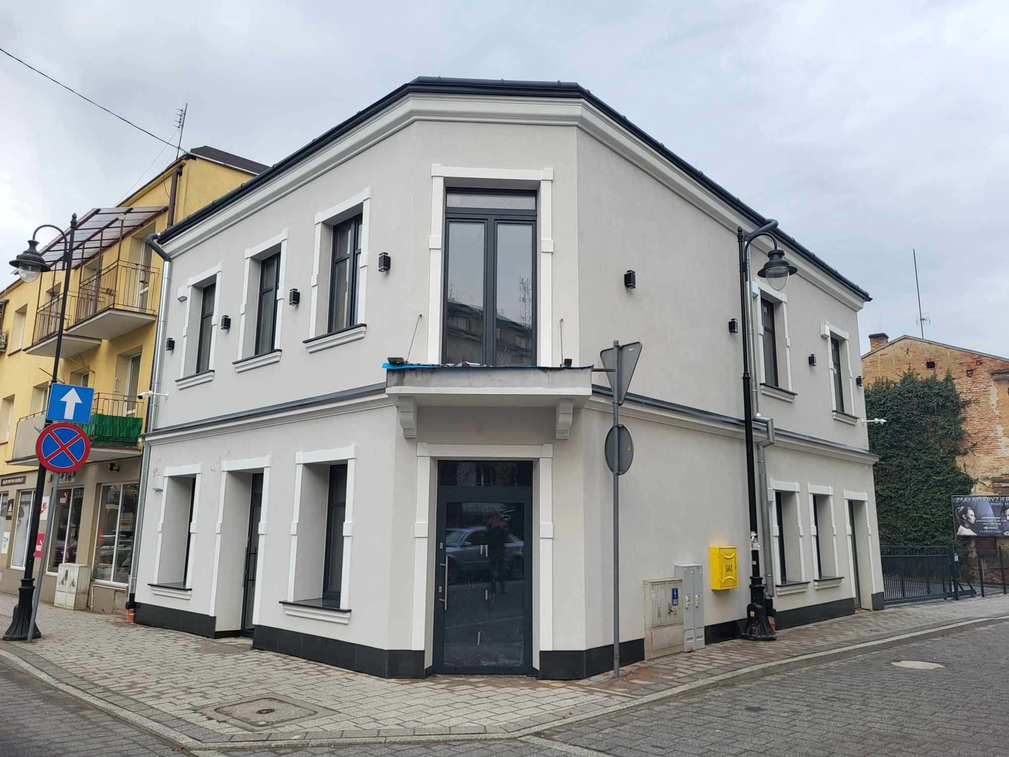 Odnowiona narożna kamienica z szarą elewacją, czarnymi ramami okiennymi i białymi obramowaniami, widok z poziomu ulicy na skrzyżowaniu, z żółtą skrzynką gazową na elewacji.