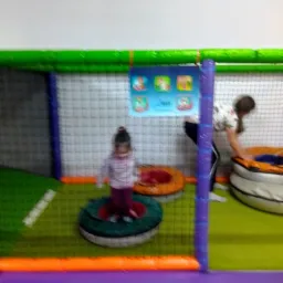 Dziewczynka stoi na okrągłej platformie w sali zabaw z siatką zabezpieczającą, w tle dorosła osoba przy basenie z kulkami.