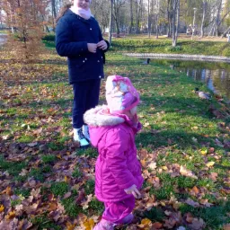 Dwie dziewczynki w parku jesienią, młodsza w r&oacute;żowym kombinezonie i czapce z postacią z bajki, starsza w granatowej kurtce i białej czapce z pomponem, w tle staw z kaczkami i drzewa.