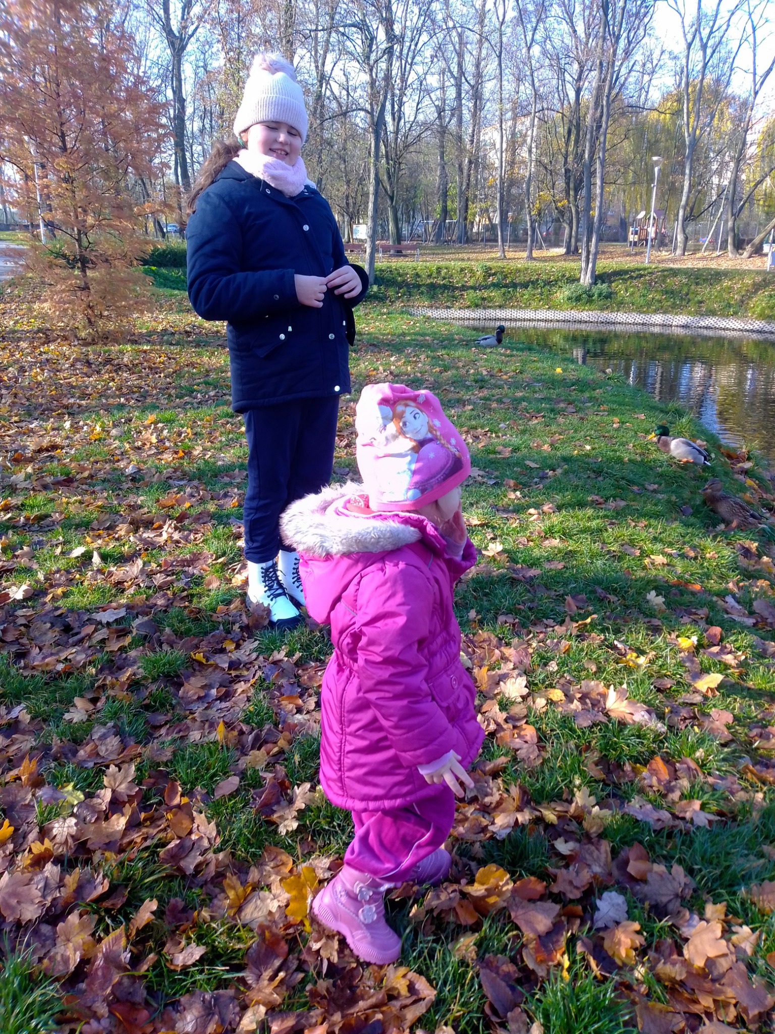 Dwie dziewczynki w parku jesienią, młodsza w różowym kombinezonie i czapce z postacią z bajki, starsza w granatowej kurtce i białej czapce z pomponem, w tle staw z kaczkami i drzewa.