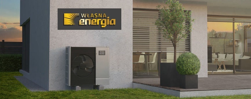 Zewnętrzna jednostka pompy ciepła Mitsubishi Ecodan zamontowana przy nowoczesnym domu z logo 'Własna Energia' na ścianie, widok na taras z meblami ogrodowymi i zachodzące słońce w tle.