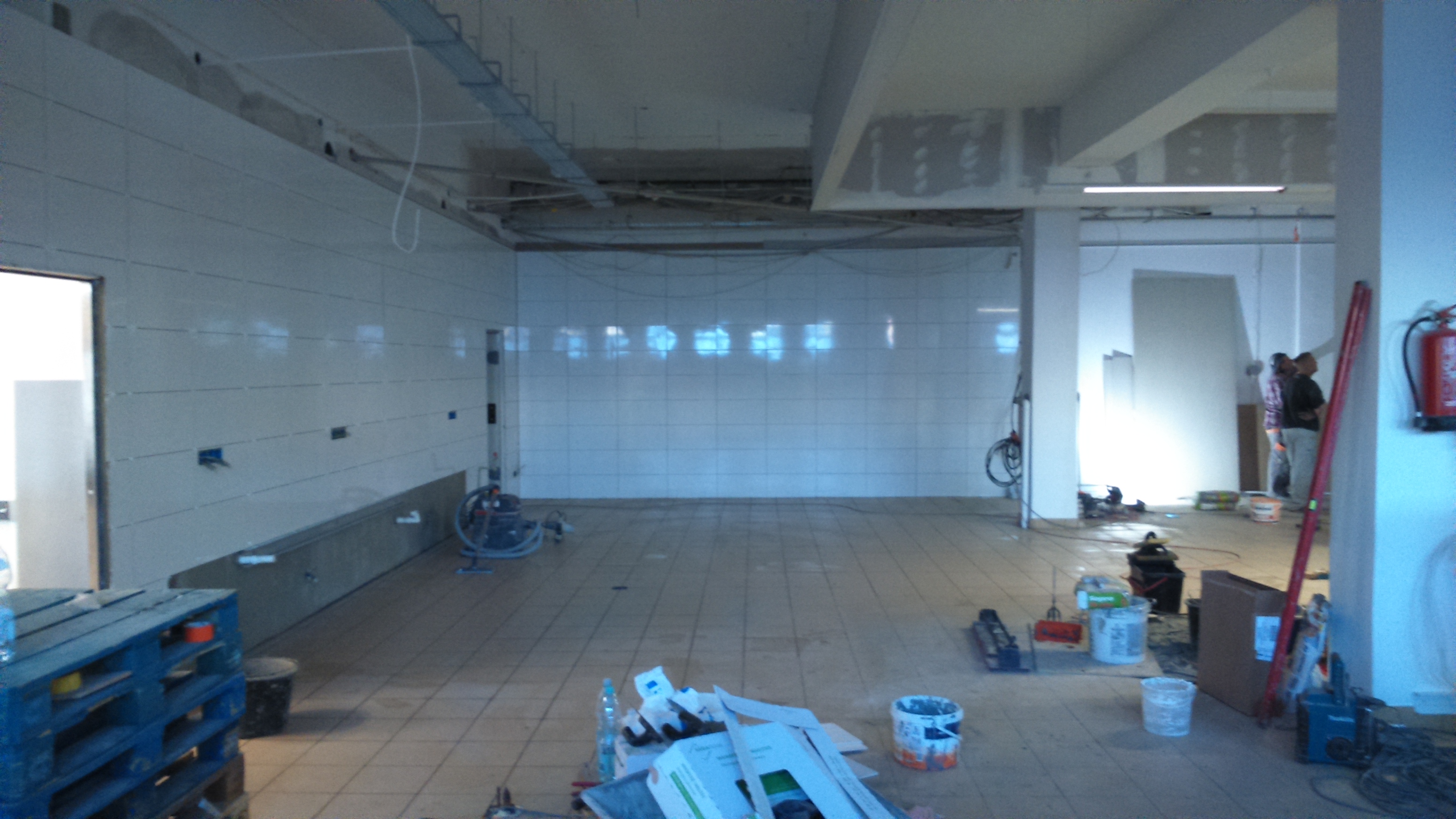 Wnętrze remontowanego pomieszczenia z białymi płytkami na ścianach, widoczne narzędzia, paleta z wodą, poziomica oparta o filar i dwóch mężczyzn w tle.