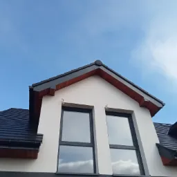 Dwuspadowy dach pokryty czarną dachówką ceramiczną, widok z dołu na lukarnę z dwoma oknami na tle błękitnego nieba z widocznym księżycem.