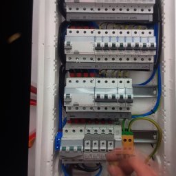 Krzysztof - Rozdzielnica elektryczna z zamontowanymi bezpiecznikami i okablowaniem, widoczne ręce osoby pracującej przy instalacji.