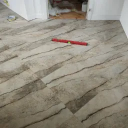 Ułożone płytki podłogowe imitujące marmur, ułożone ukośnie, z czerwoną poziomicą. W tle widoczne przygotowania do dalszych prac wykończeniowych.