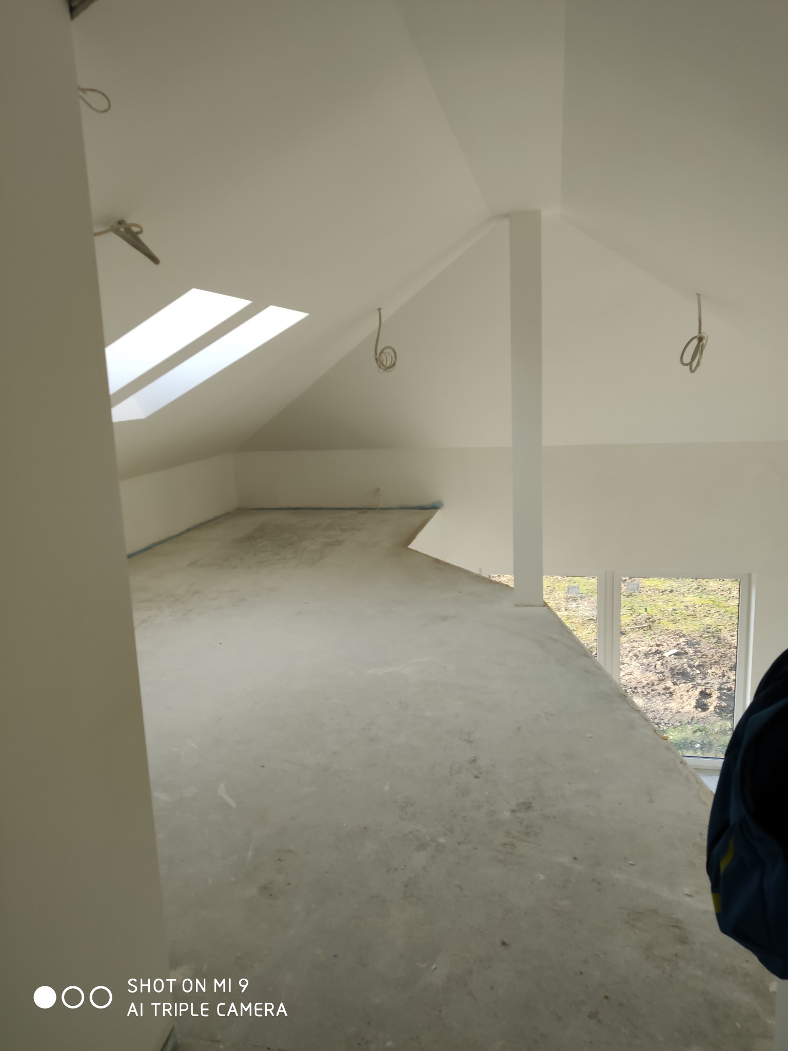 Poddasze w trakcie wykańczania, widoczne okna dachowe, niedokończona podłoga, przygotowane kable elektryczne wystające z sufitu, ściany pomalowane na biało.