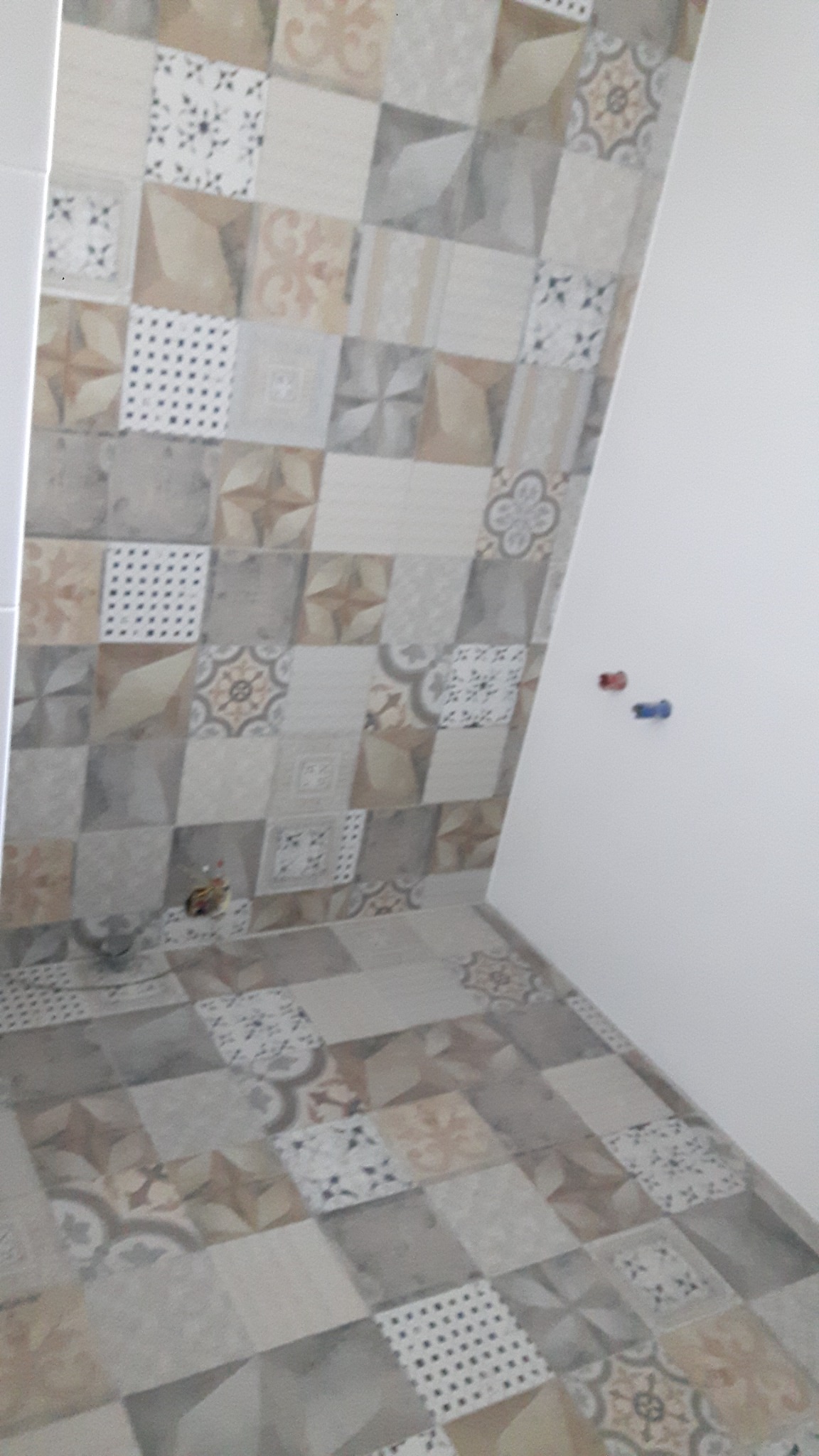 Ściana i podłoga wyłożone patchworkowymi płytkami ceramicznymi w odcieniach beżu, szarości i brązu, z widocznymi wystającymi rurami instalacji wodnej na białej ścianie.