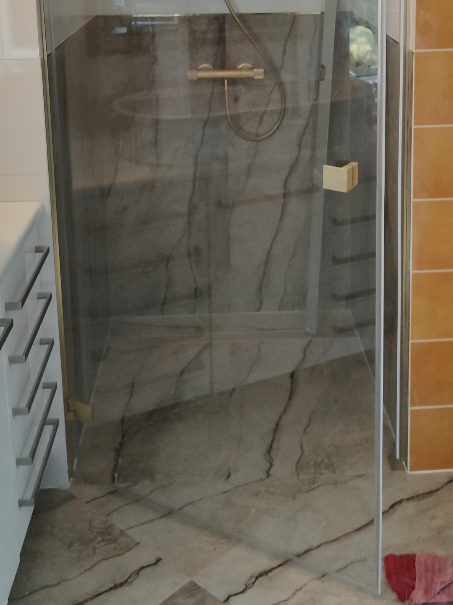 Nowoczesna kabina prysznicowa z marmurowym wykończeniem ścian i podłogi, złota armatura, widok z boku, fragment szafki z szufladami, pomarańczowe płytki na ścianie.