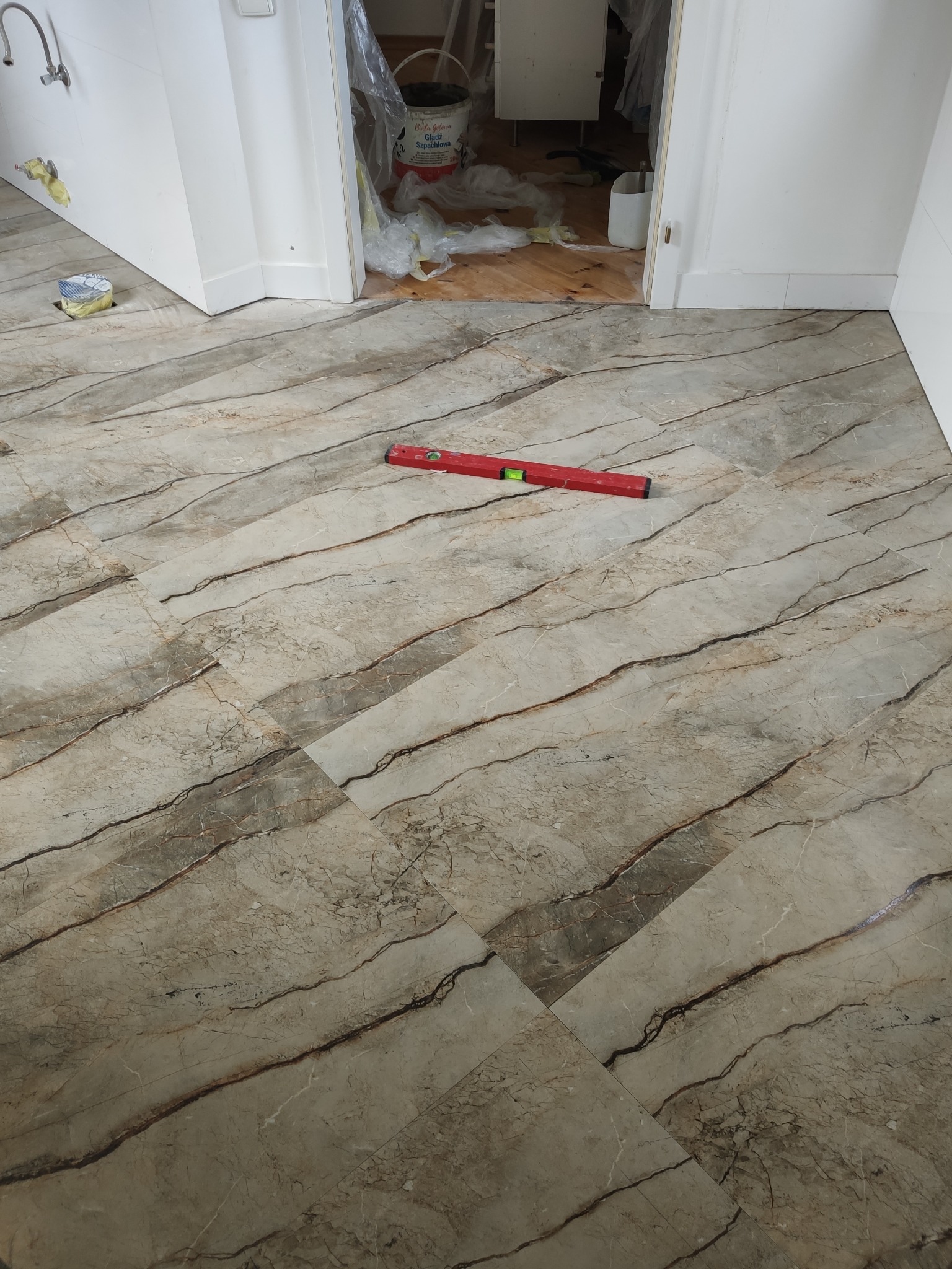 Ułożone płytki podłogowe imitujące marmur, ułożone ukośnie, z czerwoną poziomicą. W tle widoczne przygotowania do dalszych prac wykończeniowych.
