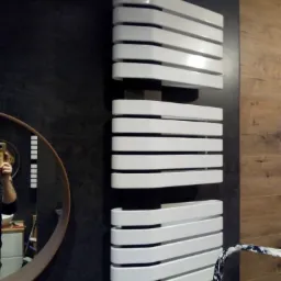 Nowoczesny, biały grzejnik łazienkowy na tle ciemnej, strukturalnej ściany, z fragmentem okrągłego lustra i drabiny malarskiej w kadrze.