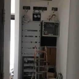 Rozdzielnia elektryczna w trakcie montażu, widoczne kable, puszki instalacyjne w ścianie, drabina malarska i narzędzia.