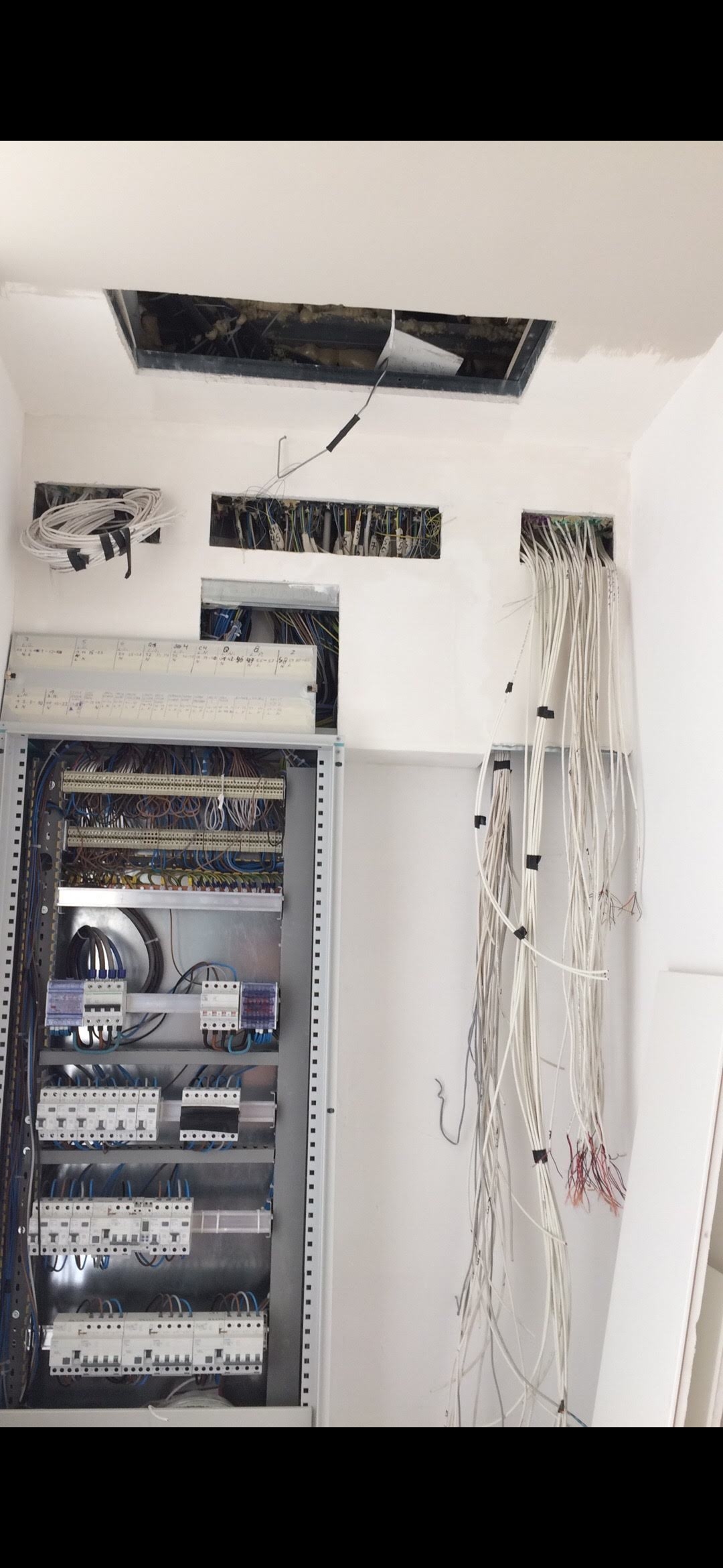 Otwarta szafa sterownicza z widocznymi zabezpieczeniami, licznymi przewodami i otworami w ścianie z przeciągniętymi kablami, w trakcie prac instalacyjnych.