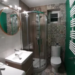 Wykończona łazienka z kabiną prysznicową, umywalką z szafką, toaletą i grzejnikiem drabinkowym; ściany pokryte płytkami w stylu patchwork oraz pomalowane na zielono.