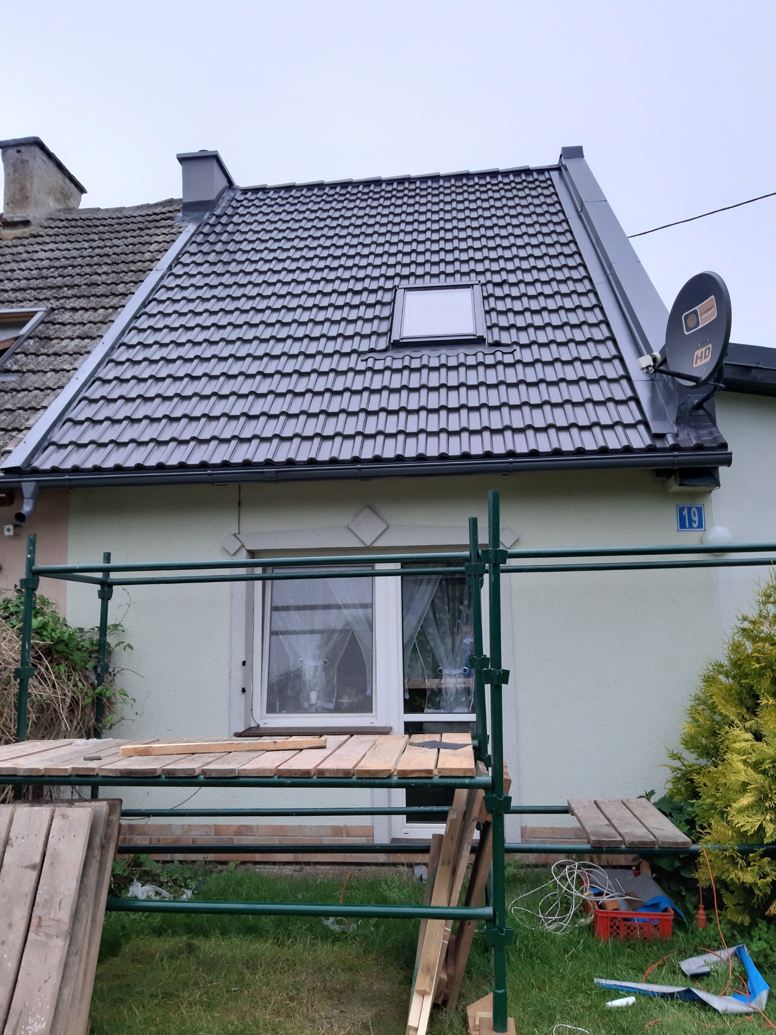 Dom z nowym, ciemnoszarym dachem z dachówki ceramicznej z oknem połaciowym i anteną satelitarną, widok z poziomu rusztowania.