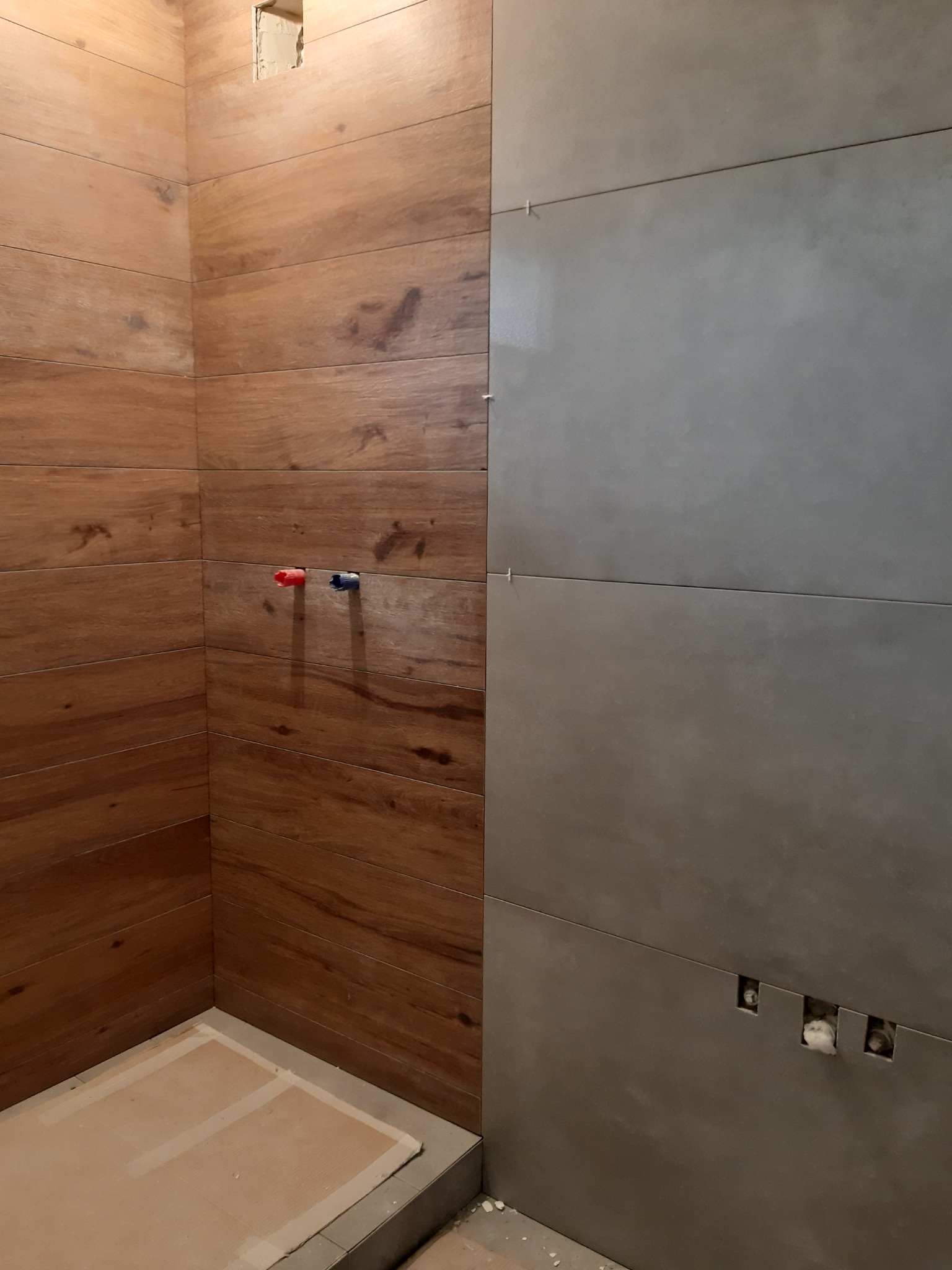 Wnętrze łazienki w trakcie wykańczania: ściany wyłożone częściowo płytkami imitującymi drewno, częściowo szarymi płytkami, widoczne otwory na instalacje wodne i elektryczne, podłoga zabezpieczona...