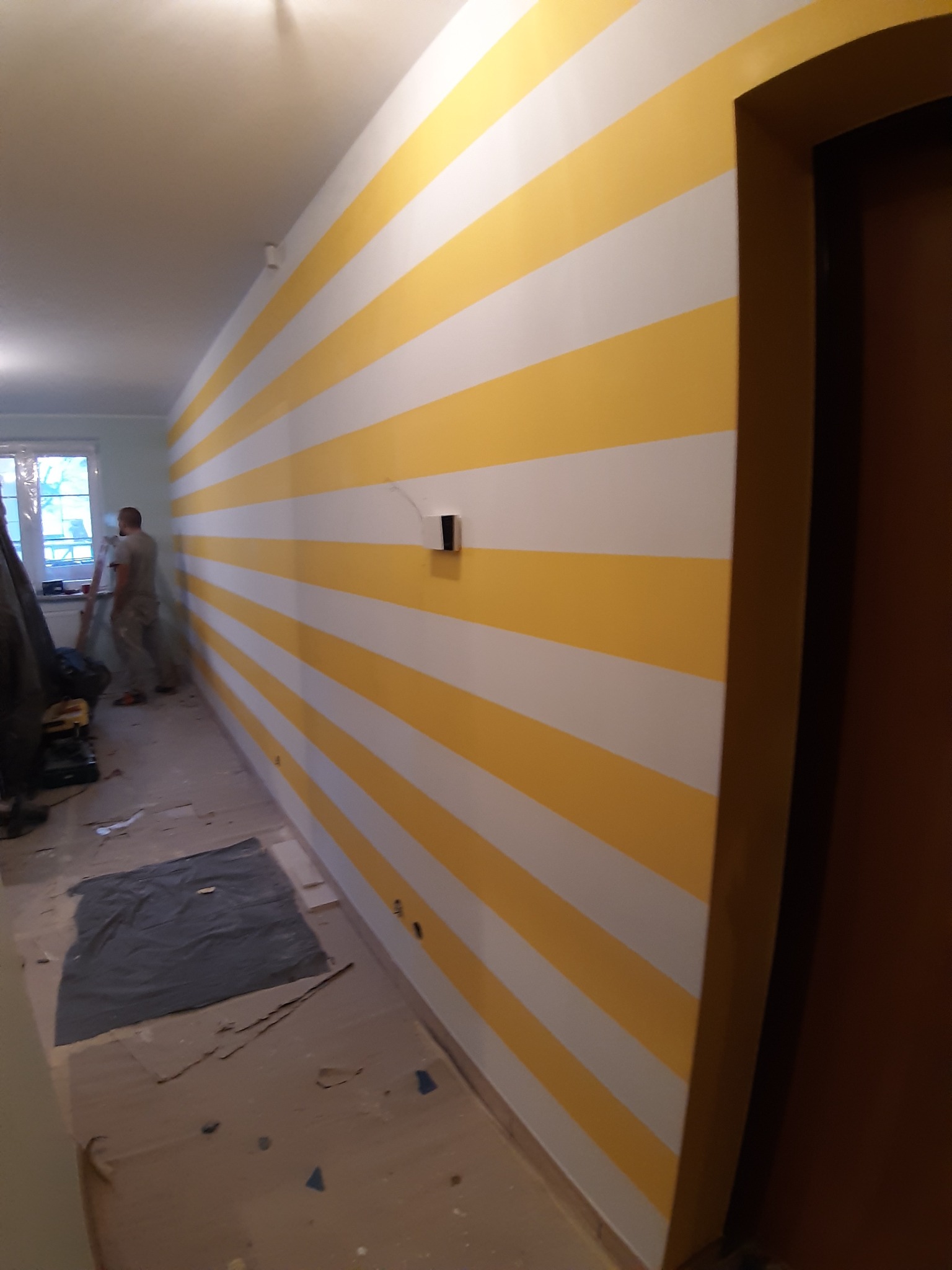 Ściana w pokoju pomalowana w poziome pasy koloru żółtego i białego, widoczny fragment futryny drzwi, podłoga zabezpieczona kartonem i folią malarską, w tle pracownik malujący ścianę przy oknie.