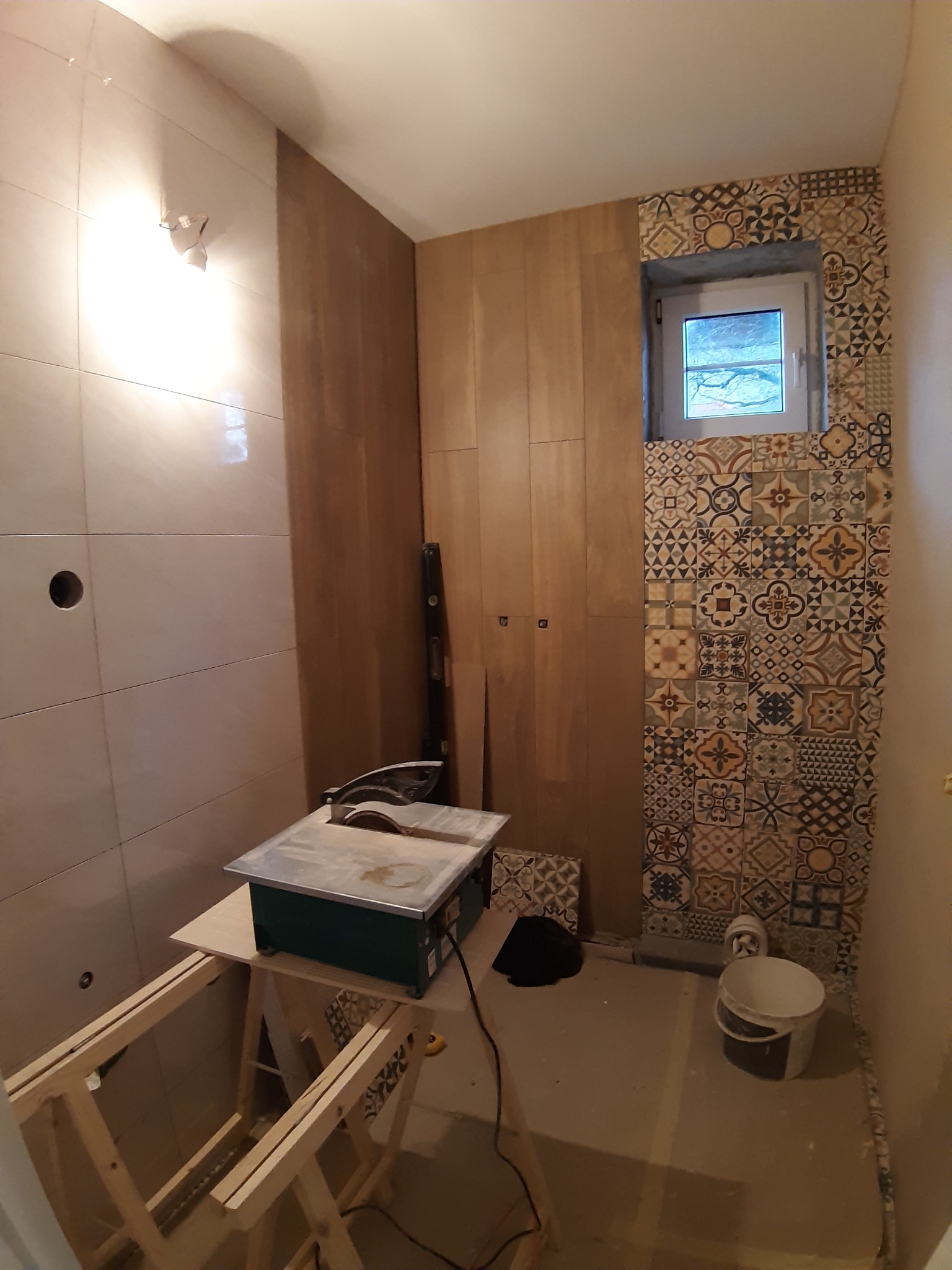 Wnętrze łazienki w trakcie prac wykończeniowych: częściowo ułożone płytki ceramiczne o zróżnicowanych wzorach, widoczna przecinarka do płytek na drewnianym stojaku, poziomica oparta o ścianę,...