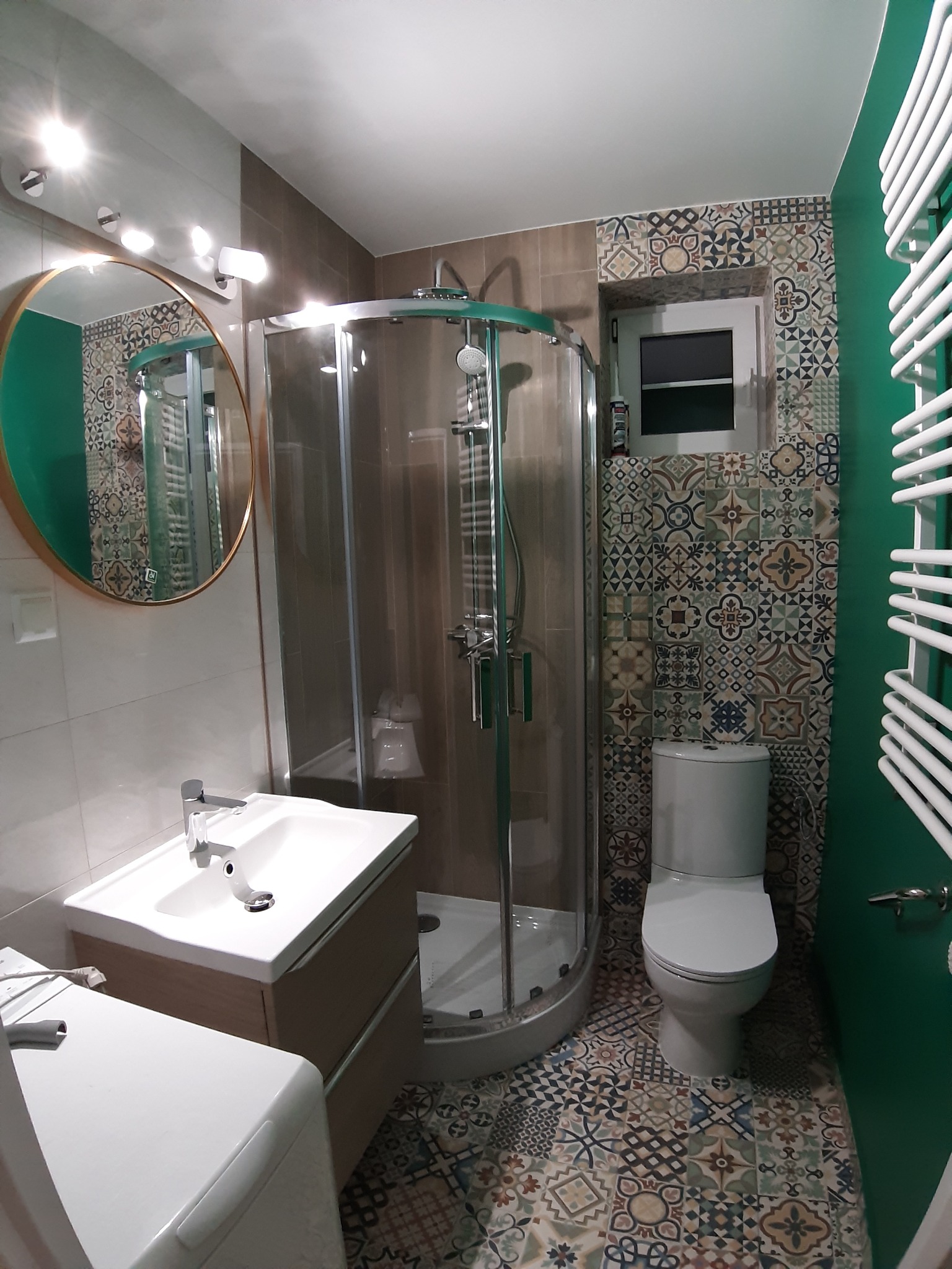 Wykończona łazienka z kabiną prysznicową, umywalką z szafką, toaletą i grzejnikiem drabinkowym; ściany pokryte płytkami w stylu patchwork oraz pomalowane na zielono.