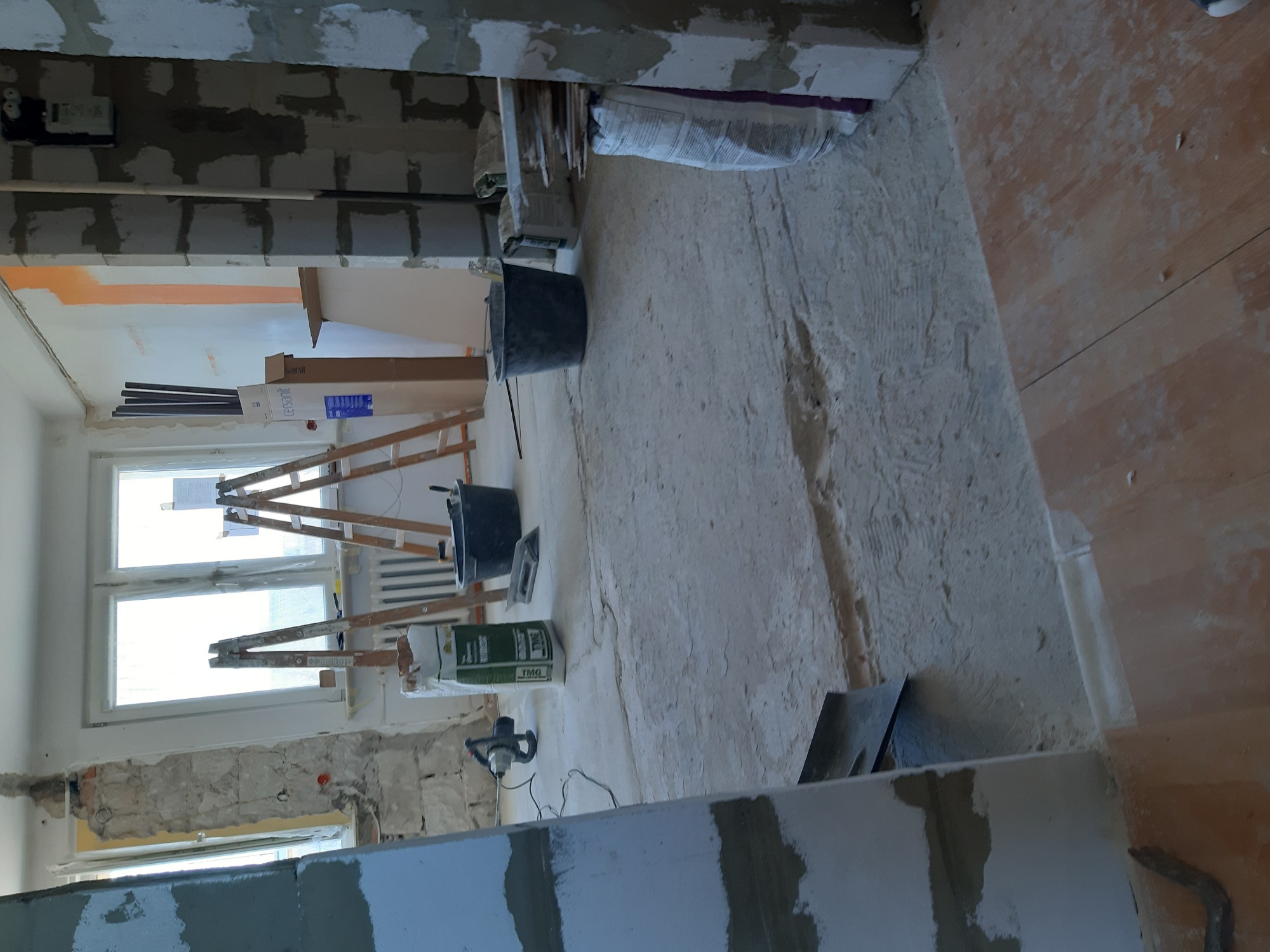 Wnętrze w trakcie prac w Elblągu: widok na pokój z częściowo oszlifowaną podłogą, ściany w trakcie przygotowania, widoczne drabiny, wiadra, worki z zaprawą i mieszadło elektryczne.