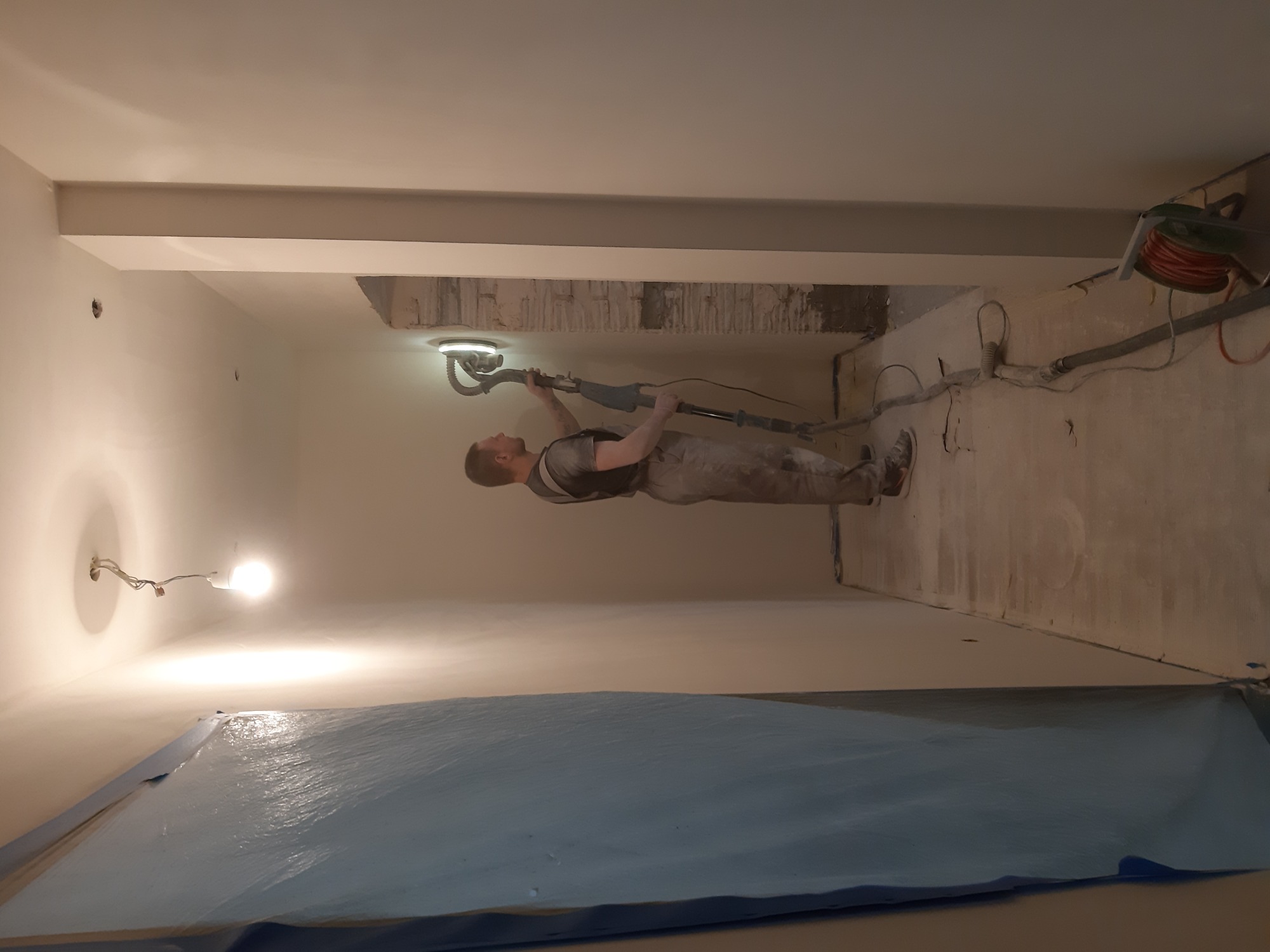 Pracownik szlifuje ścianę w pomieszczeniu za pomocą szlifierki z odkurzaczem; podłoga zabezpieczona folią ochronną, widoczne przewody i instalacje.