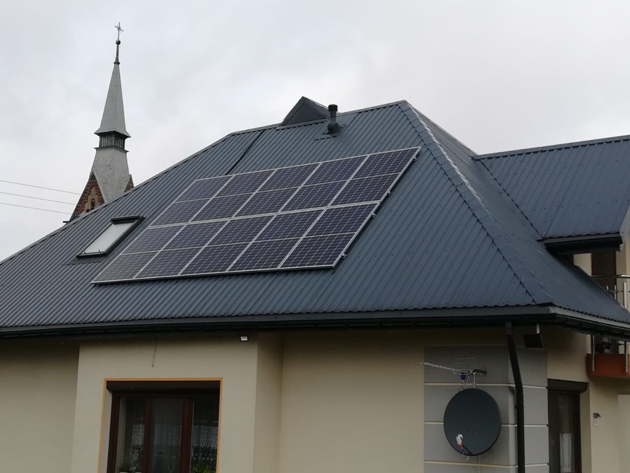 Instalacja paneli słonecznych na dachu domu jednorodzinnego z blachy trapezowej, widoczny również kościół w tle.