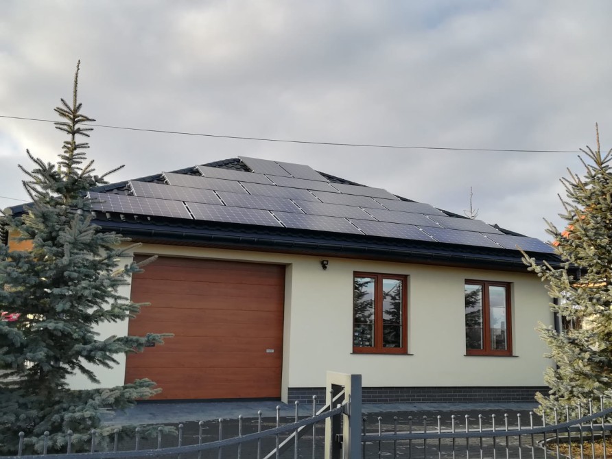 Instalacja fotowoltaiczna na dachu domu jednorodzinnego z brązową bramą garażową i dwoma świerkami kłującymi po bokach, widok zza metalowego ogrodzenia.