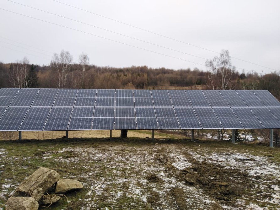 Instalacja fotowoltaiczna na gruncie w okolicy Krosna, widoczne panele na metalowej konstrukcji, w tle drzewa i pochmurne niebo.