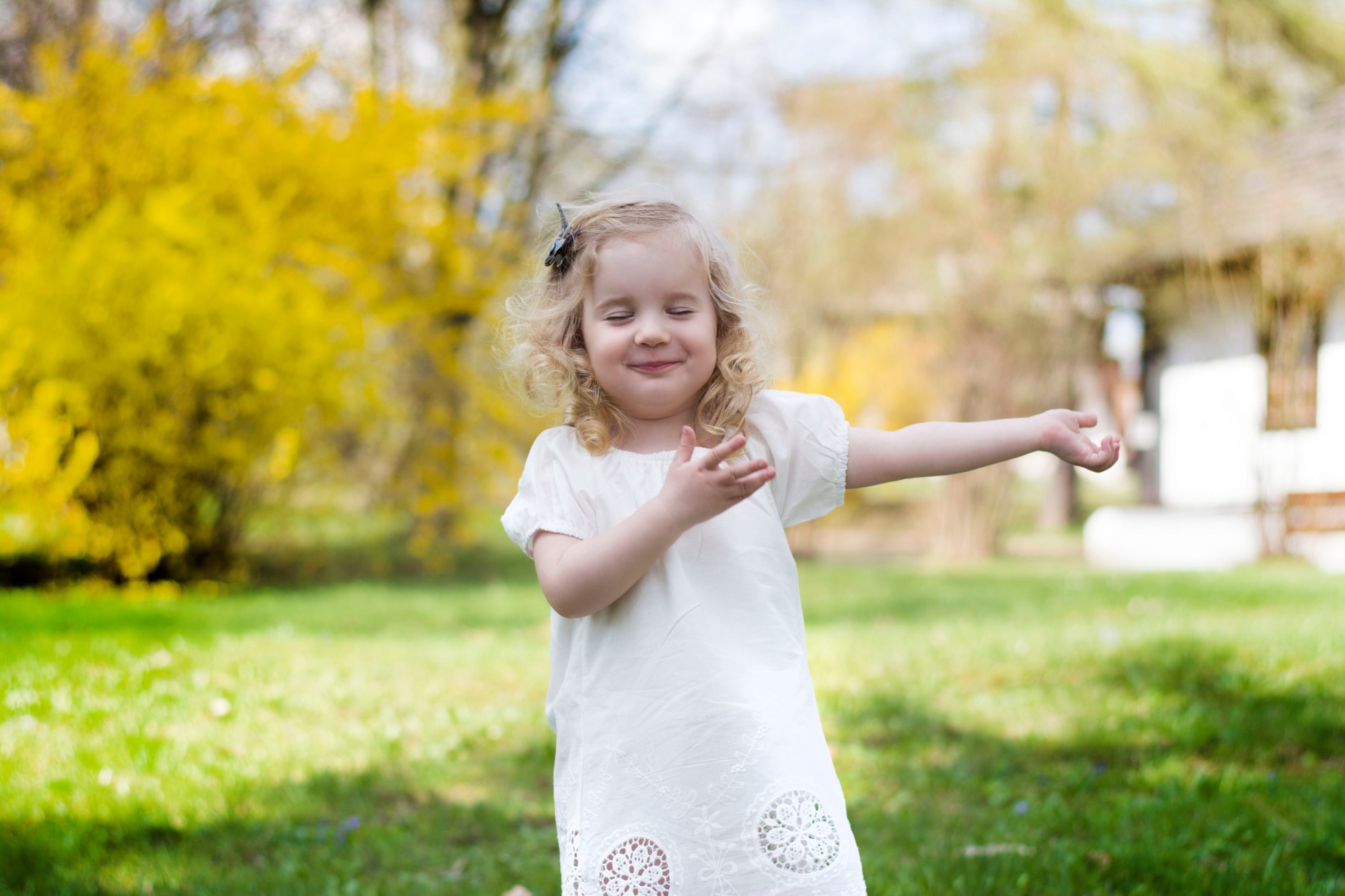 Uśmiechnięta dziewczynka w białej sukience z koronkowymi detalami, z zamkniętymi oczami, pozuje na tle kwitnących, żółtych krzewów i zielonego trawnika.